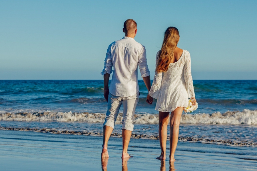 A bride and groom in casual attire walking along the beach.